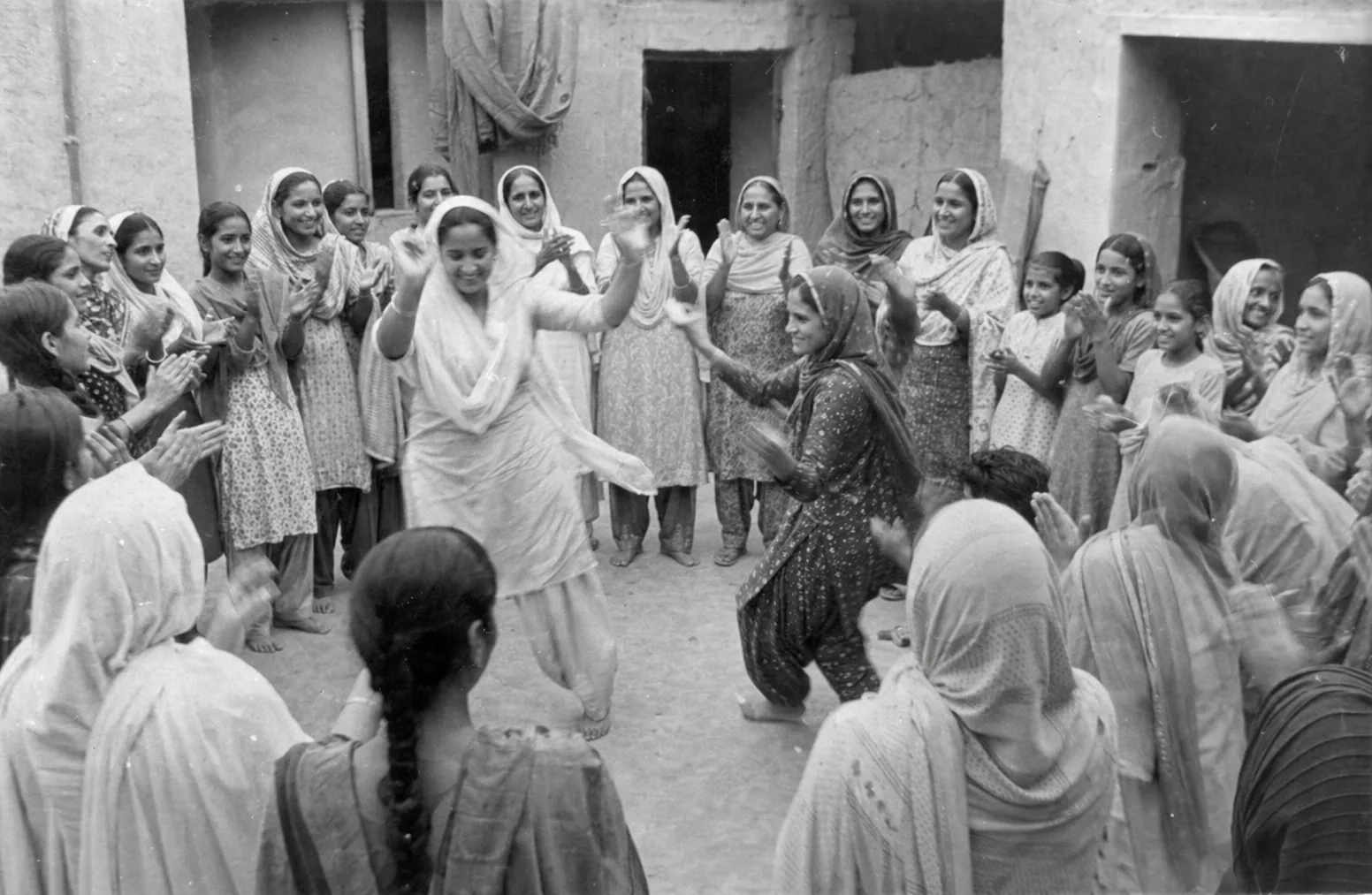 Punjabi women in a gidha circle, clapping, one leaning in to sing a boli